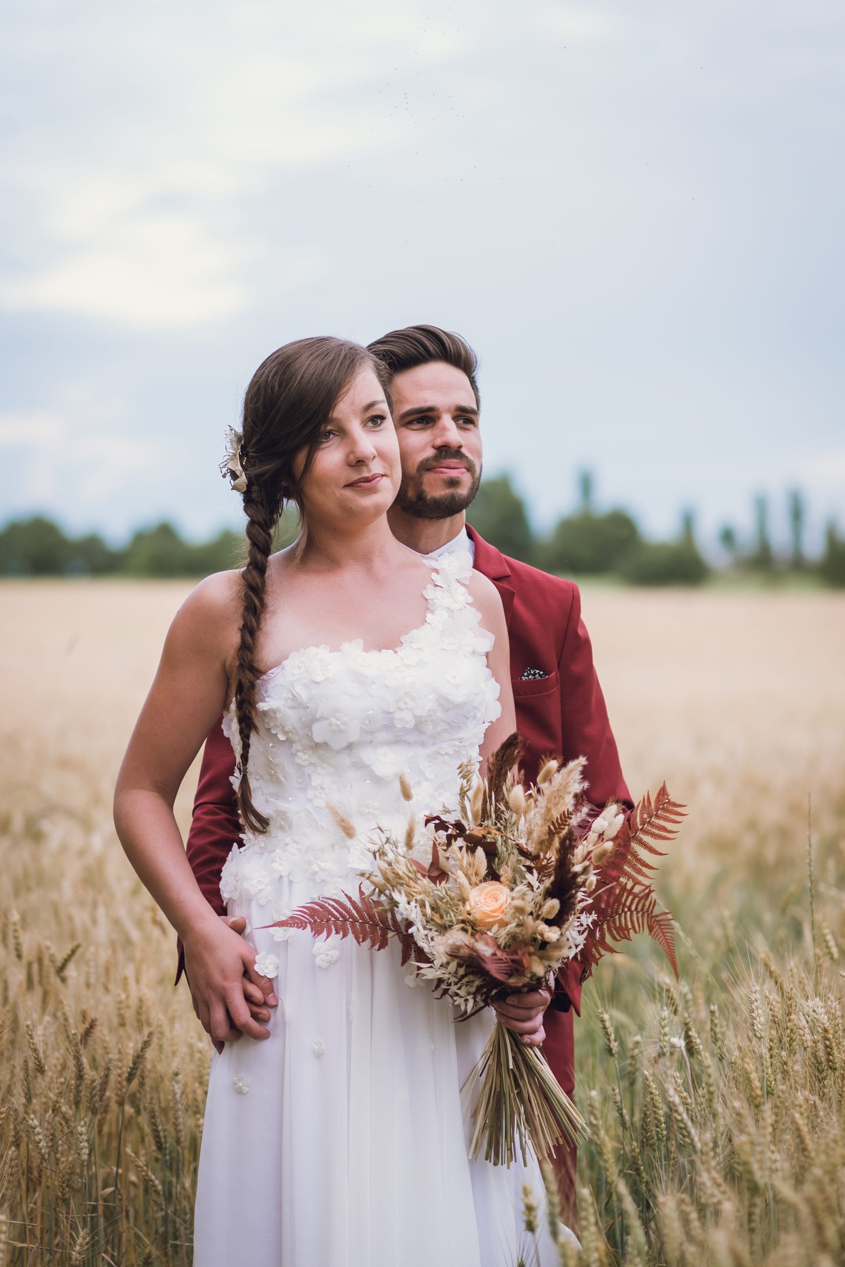Un couple photographié dans un champ de blé, la mariée tenant un bouquet de fleurs séchées, tandis que le marié porte un costume rouge, créant une scène champêtre et élégante.